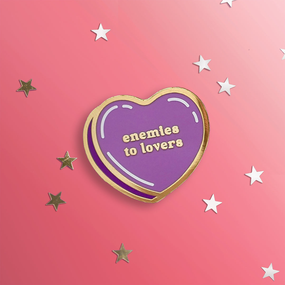 Purple heart pin with gold outline: enemies to lovers shown on a pink background with stars.
