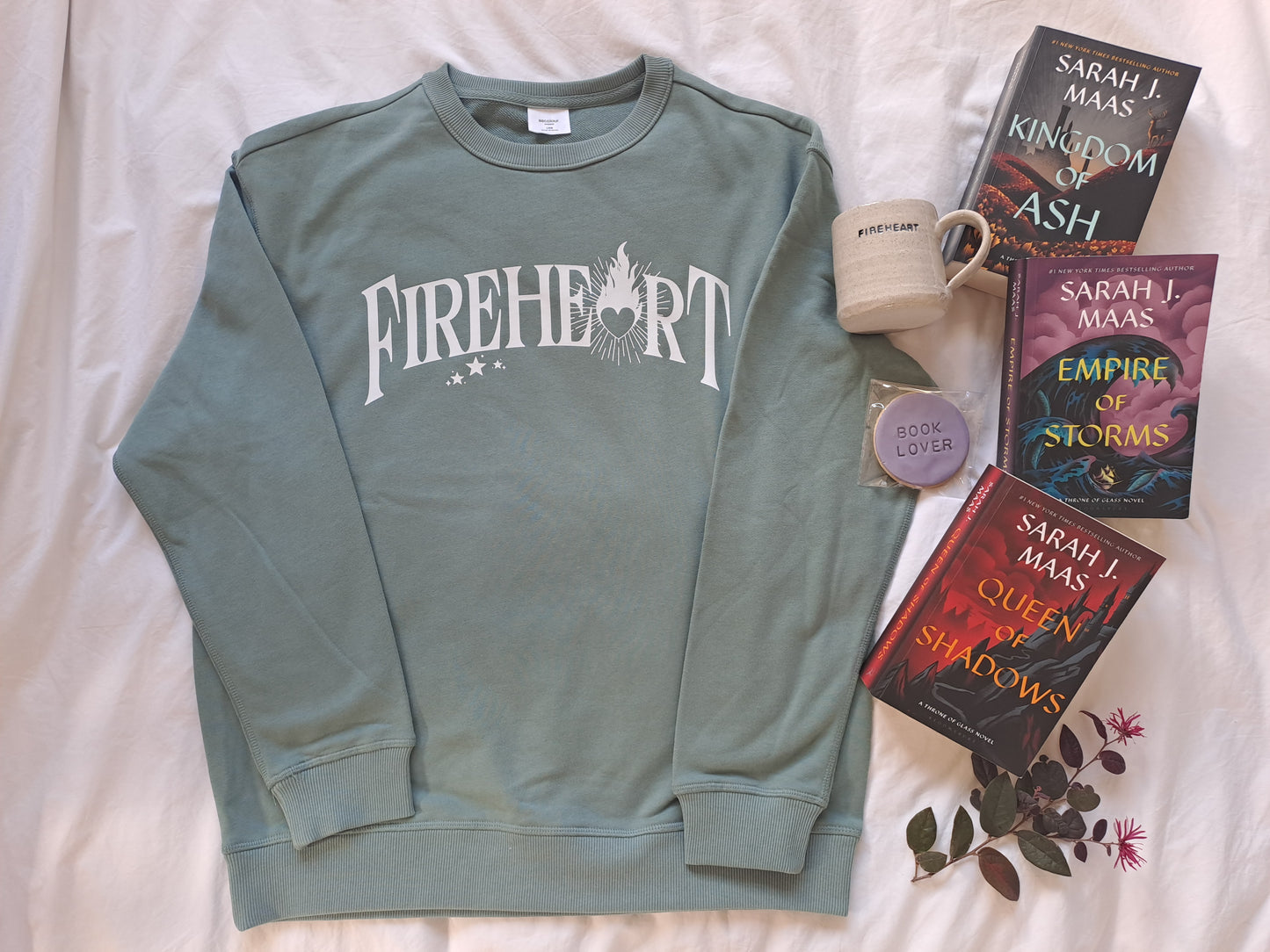 A sage green folded sweatshirt with white text "Fireheart". The A is replaced with a flaming heart. Shown as a flatlay with tea, Fireheart mug and Throne of Glass books by Sarah J Maas.