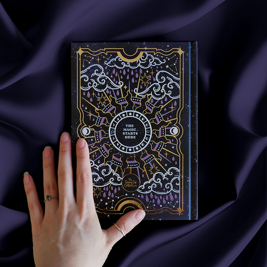 Black journal with intricate gold and purple patterns, shown on a purple satin background with a hand on top.
