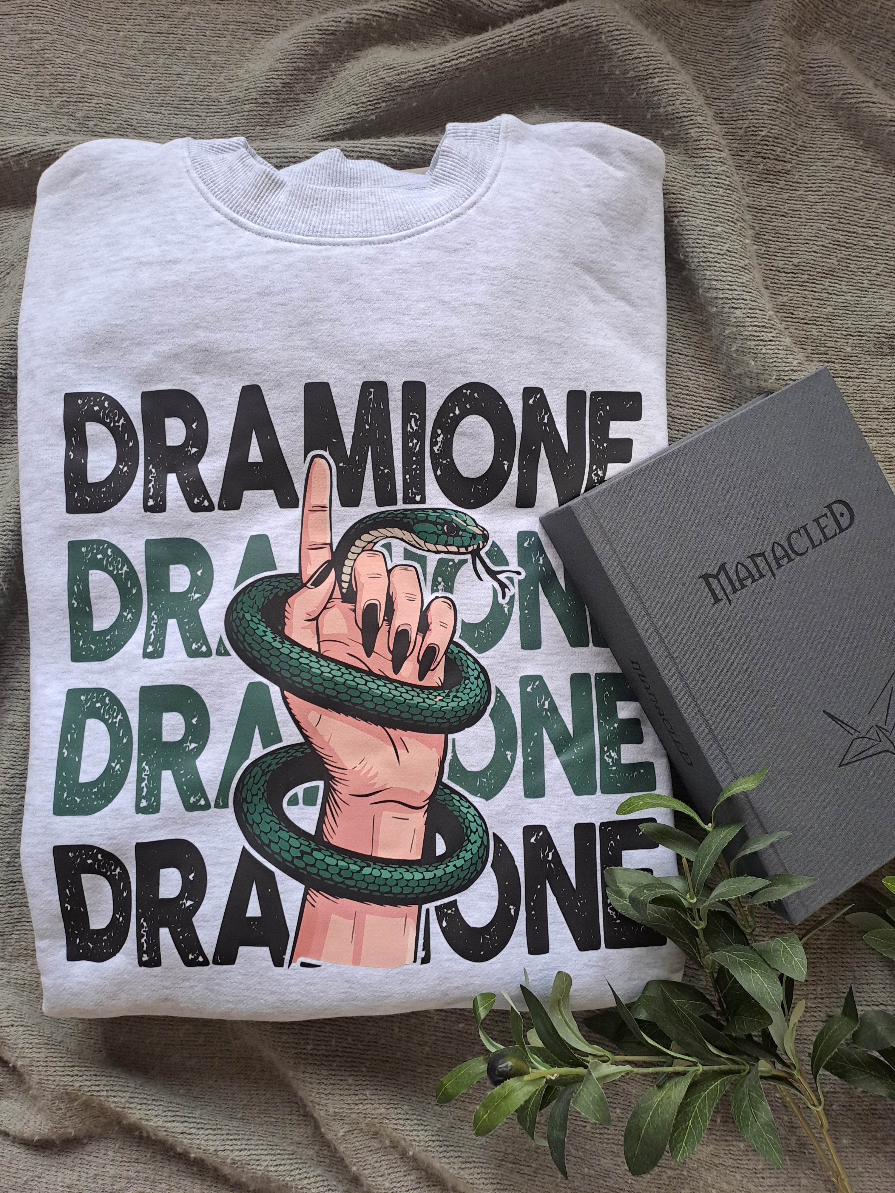 A light grey folded sweatshirt with an image of a female hand wrapped in a snake and "Dramione" text behind it. Shown as a flatlay with the epic fan fiction novel Manacled.