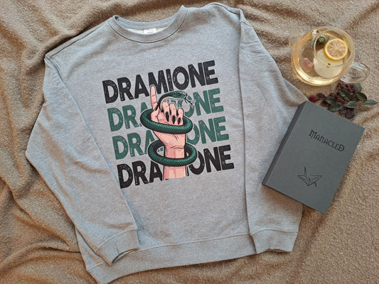 A grey sweatshirt with an image of a female hand wrapped in a snake and "Dramione" text behind it. Shown as a flatlay with tea and the epic fan fiction novel Manacled.
