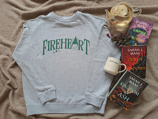 A grey folded sweatshirt with green text "Fireheart". The A is replaced with a flaming heart. Shown as a flatlay with tea, Fireheart mug and Throne of Glass books by Sarah J Maas.