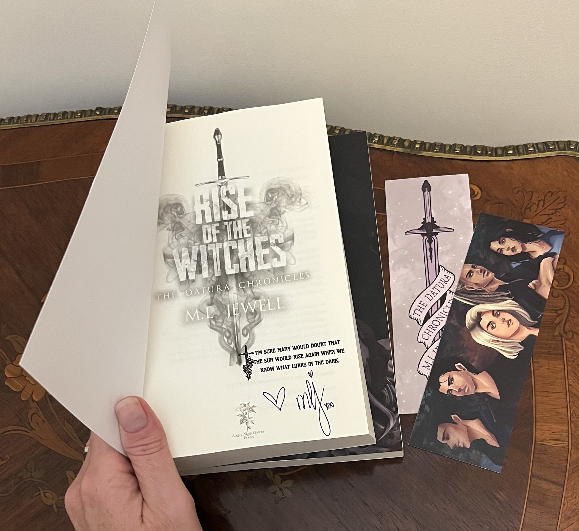 Book titled 'Rise of the Witches' by M.L. Jewel held open to see the author's signature and matching bookmark.