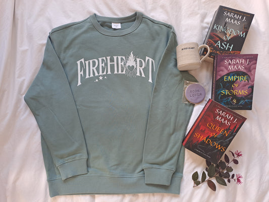 A sage green folded sweatshirt with white text "Fireheart". The A is replaced with a flaming heart. Shown as a flatlay with tea, Fireheart mug and Throne of Glass books by Sarah J Maas.