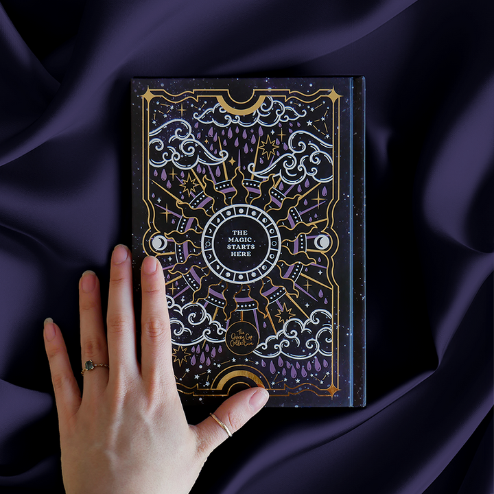 Black journal with intricate gold and purple patterns, shown on a purple satin background with a hand on top.