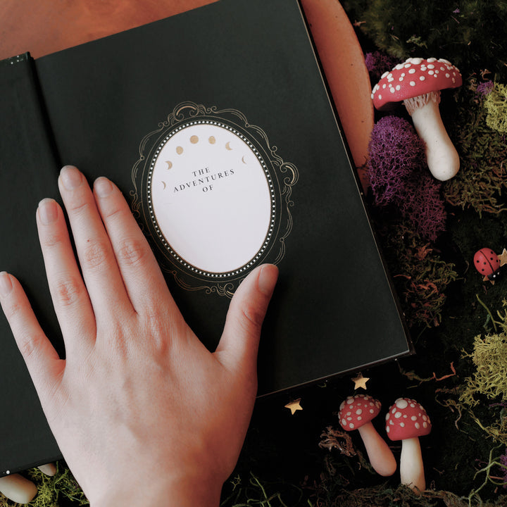 Dark green journal open to first page,  shown on a forest fantasy flat lay with a hand on top.