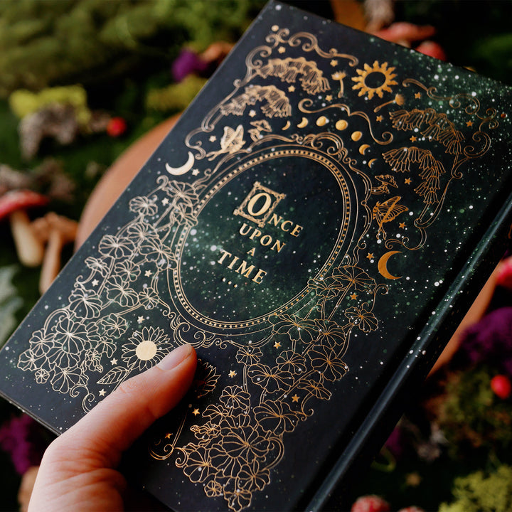 Dark green journal with intricate gold patterns, shown on a forest fantasy flat lay with a hand on top. Showing the back which says Once Upon A Time.